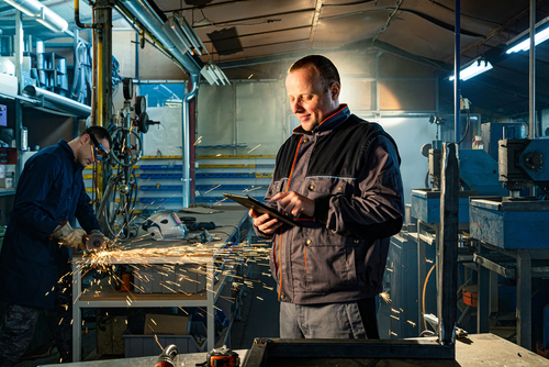 Fabricator reviewing job on tablet while colleague grinds steel in the background