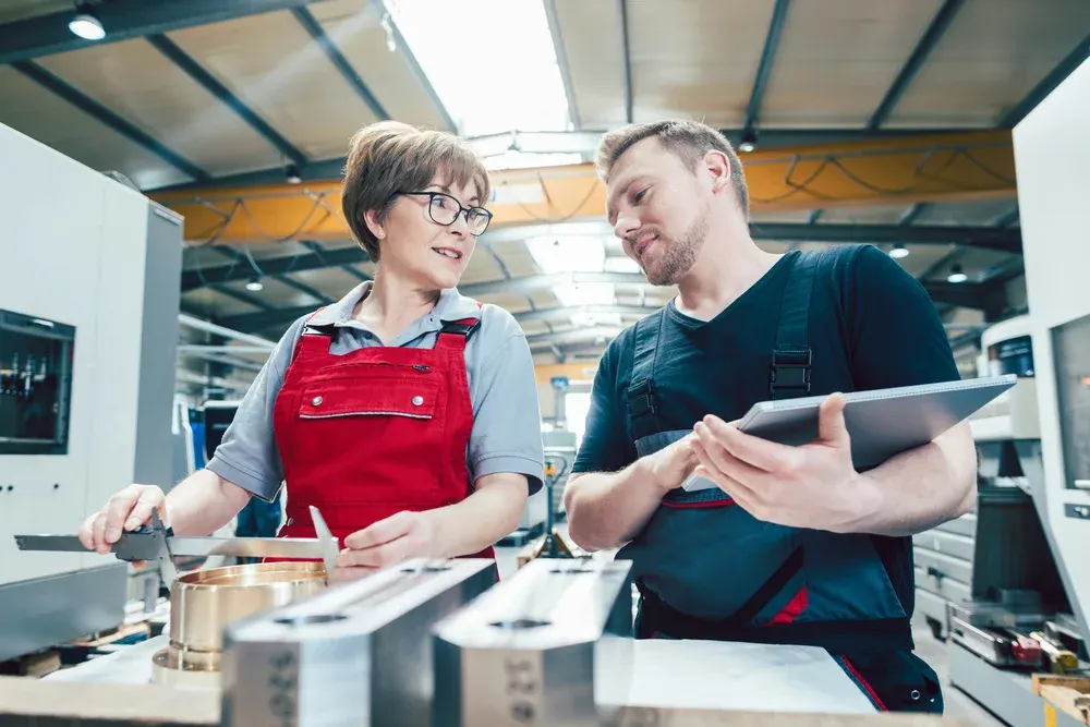 Two manufacturing team members inspecting a component with calipers and a tablet